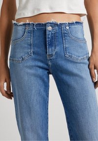 Höga blå denimjeans med fransad nederkant, fem fickor och framknäppning. Tyget har en något strukturerad yta.