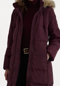 Burgundské péřové sako se zipem a patentním uzávěrem, vybavené kapucí podšitou umělou kožešinou. Látka má hladký, matný povrch a je prošívaná.