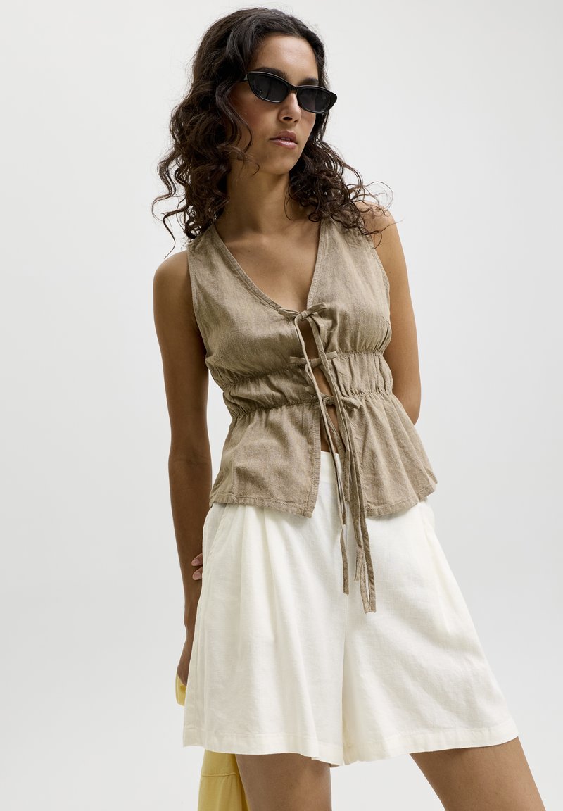 Femme aux cheveux bouclés portant des lunettes de soleil noires, un haut beige sans manches noué devant et un short blanc à jambes larges sur un fond uni.