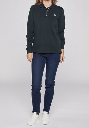 Femme portant un polo noir à manches longues, un jean bleu foncé et des baskets blanches, debout devant un fond clair uni.