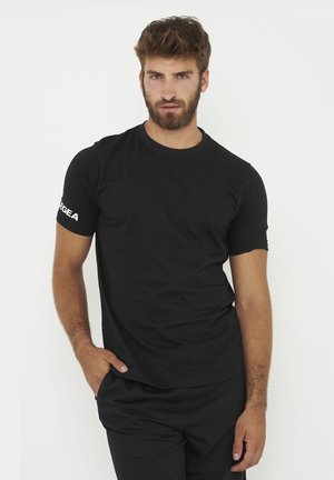 Uomo con la barba che indossa una maglietta e pantaloni neri, in piedi con una mano in tasca su sfondo bianco semplice.