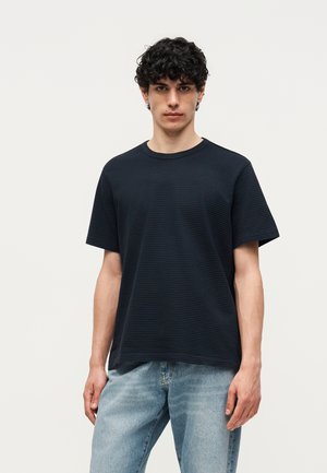 Νέος άνδρας με σγουρά μαλλιά, που φοράει μαύρο υφασμάτινο κοντομάνικο πουκάμισο και ανοιχτό μπλε τζιν, στέκεται μπροστά σε μια απλή φωτεινή επιφάνεια φόντου.