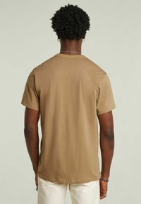 T-shirt in cotone marrone, collo rotondo, maniche corte, texture liscia, forma aderente, dettagli minimi, indossato con pantaloni chiari.