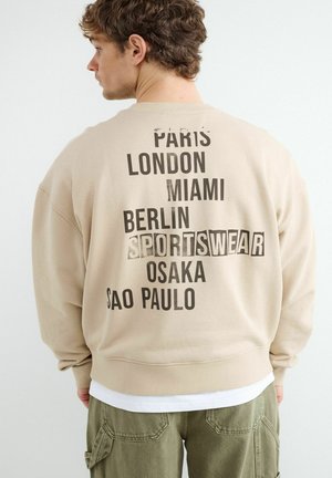 Jeune homme de dos portant un sweat-shirt beige avec les noms des villes Paris, Londres, Miami, Berlin, Osaka, Sao Paulo et le mot « Sportswear » dans le dos.