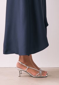 Ezüstszínű, vékony pántos magassarkú szandálok, finom boka csattal és karcsú, 3 hüvelyk magas sarokkal, egy légies, sötétkék ruhával párosítva.