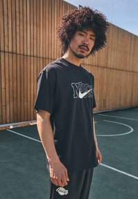 Černé bavlněné tričko s výrazným bílým logem Nike. Spárováno s černými kalhotami, které zobrazují bílé logo Lakers. Venku ve sportovním prostředí.