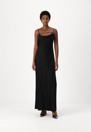Mustahline musta maxi dress dengan tali tipis dan bodice yang pas; menampilkan sedikit kilau, desain asimetris, dan tekstur yang halus. Sepatu hak tinggi hitam.