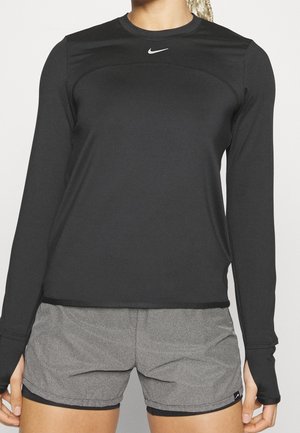 Femme portant un haut de sport noir à manches longues avec des trous pour les pouces et un short de course gris texturé sur fond uni.