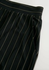 Pantalon en tissu noir avec fines rayures dorées verticales, taille élastique et poches latérales. Texture douce avec une coupe ajustée.