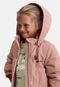 Rosa Jacke mit Reißverschluss und Kapuze aus leichtem Stoff. Verziert mit Stickereien und einem kontrastierenden Innenfutter. Wird über einem grünen Shirt getragen.