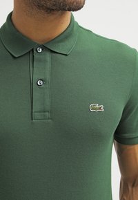 Zelené polo tričko vyrobené z texturované tkaniny s límečkem, které má zapínání na tři knoflíky a bílou logo krokodýla na hrudi.