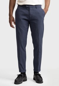 Pantalons tailoring bleu marine avec une texture lisse, coupe slim, deux poches latérales et une taille mi-haute. Portés avec des chaussures et des chaussettes noires.