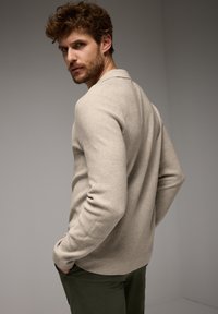 Béžový pletený svetr s texturovaným povrchem, s límečkem a dlouhými rukávy, doplněný tmavě zelenými kalhotami.