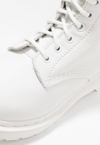 Stivale bianco in pelle con lacci bianchi infilati attraverso occhielli metallici, cuciture dettagliate e una suola in gomma testurizzata su uno sfondo bianco.