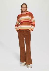 Gestreepte gebreide trui in oranje, beige en roze tinten, gecombineerd met bruine corduroy broek en beige plateauschoenen.