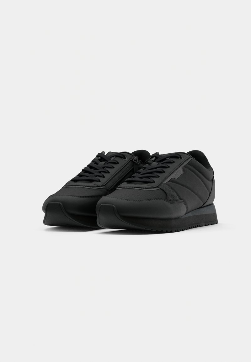 Une paire de baskets de sport noires avec des lacets et un détail de fermeture éclair sur le côté, présentée de l'angle frontal sur un fond blanc.