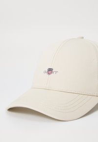 Beige baseballpet van canvas met een gebogen rand. Voorzien van een geborduurd logo op de voorkant en stikselaccenten.