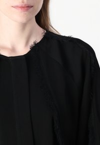 Black garment with a simple design, featuring textured fringe along the neckline and shoulders; smooth fabric with a matte finish.