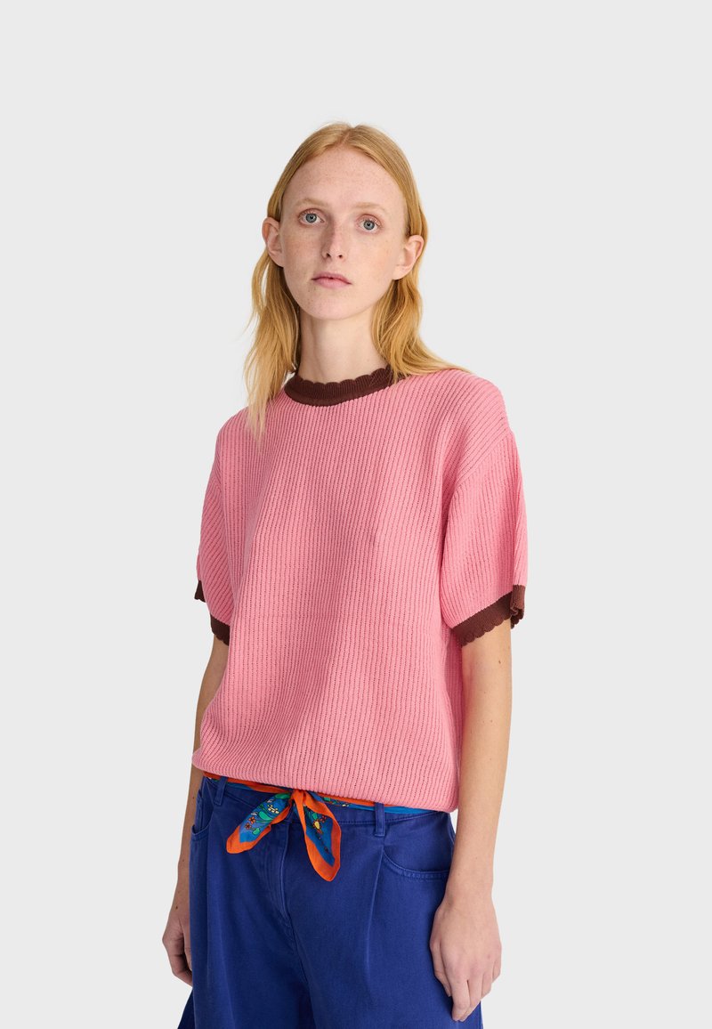Jeune personne aux cheveux rouges et raides portant un haut rose en maille à manches courtes avec une bordure marron et un pantalon bleu avec une ceinture orange à motifs.