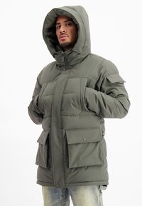Man in een groene gewatteerde hoodie met grote klepzakken en rits, met zijn handen in de zakken, tegen een witte achtergrond.