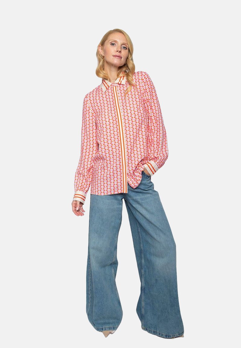 Rote und rosa gemusterte Bluse mit Kragen, langen Ärmeln und orangefarbenen Akzenten, kombiniert mit weit geschnittenen blauen Jeans. Strukturierten Stoff.