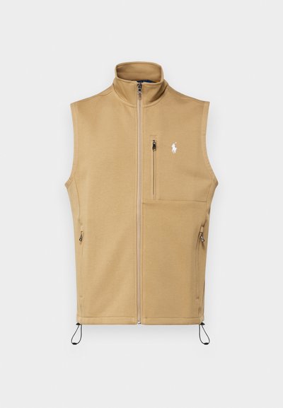 Beige glidelåsvest laget av myk stoff, med høy krage, sidelommer og et brodert logo på brystet. Justerbar snor i bunnen.