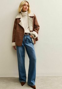Giacca in shearling marrone con rivestimento crema, abbinata a un dolcevita beige, jeans a gamba larga blu e scarpe con punta a cono marroni.