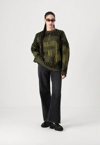 Πουλόβερ υπερμεγέθες πράσινο πλεκτό με υφασμένα μοτίβα, σε συνδυασμό με σκουρόχρωμα τζιν με φαρδιά πόδια και μαύρα παπούτσια με πλατφόρμα. Το μοντέλο φοράει μαύρα γυαλιά ηλίου.