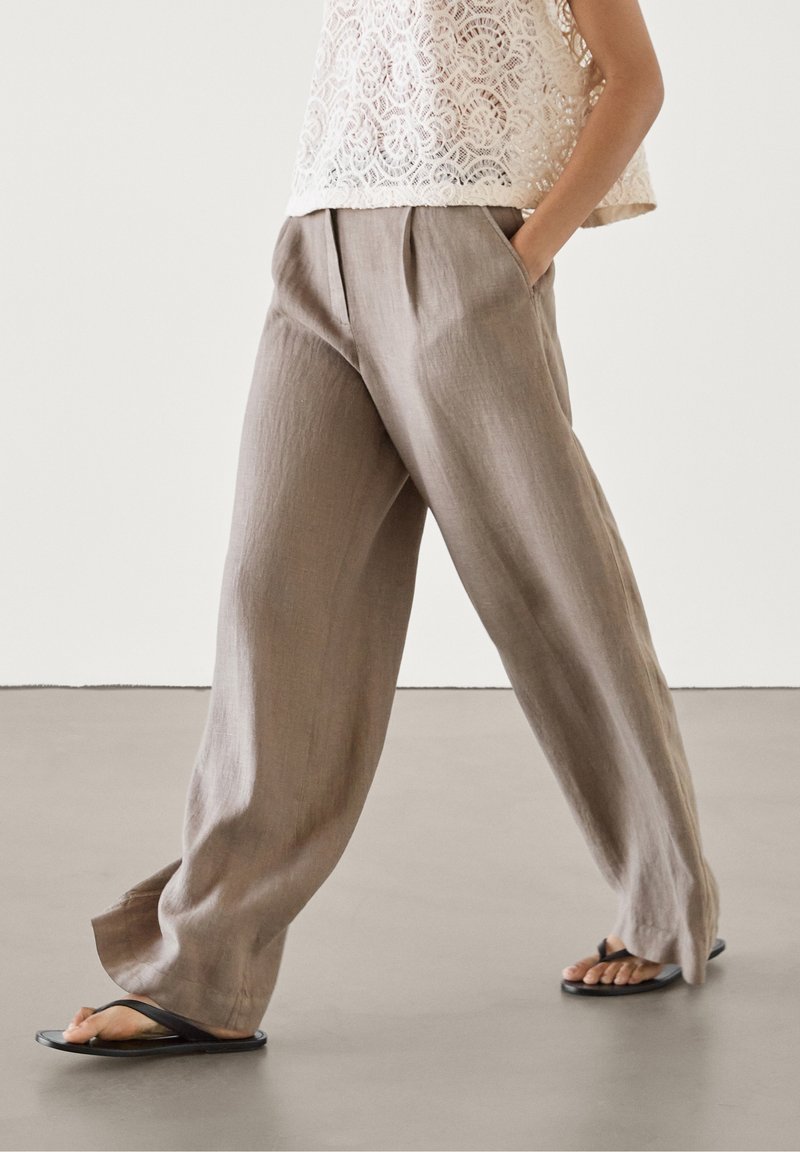 Pantalon large en lin marron clair avec plis et poches latérales, associé à un haut en dentelle crème et des tongs noires. Texture lisse.