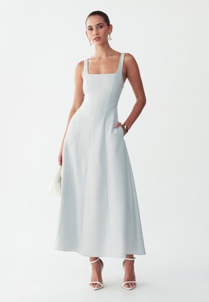 Vrouw draagt een lichtblauwe mouwloze midi-jurk met een vierkante halslijn, witte hoge hakken sandalen, houdt een kleine witte handtas vast.