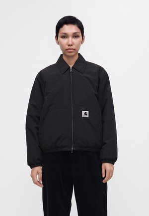 Person med kort mørkt hår iført en sort lynlåstrøje med et lille hvidt Carhartt logo på lommen, stående foran en ensfarvet baggrund.
