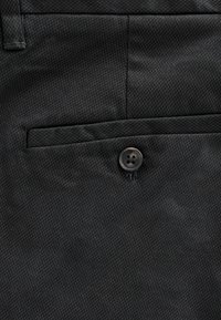 Pantaloni din țesătură neagră cu o suprafață texturată, o buzunare frontală cu închidere prin buton și cusături îngrijite pe marginile acestora.