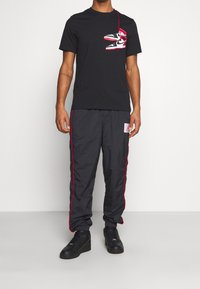 Camiseta negra con un gráfico de zapatillas rojas y blancas, combinada con pantalones de nailon negros con franjas laterales rojas y un parche. Zapatillas negras completan el look.