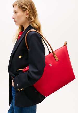 Vrouw in een marineblauwe blazer en spijkerbroek draagt een grote rode boodschappentas met bruine handvatten over haar schouder tegen een witte achtergrond.