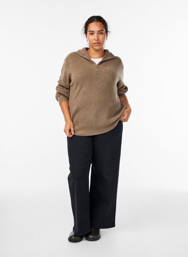 Woman with dark hair wearing a brown knit sweater, black wide-leg pants, and black shoes standing against a white background.