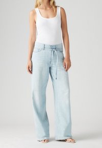 Mujer vestida con una blusa sin mangas blanca y unos pantalones de tiro alto de pierna ancha en azul claro con un cinturón de lazo y sandalias de punta abierta.