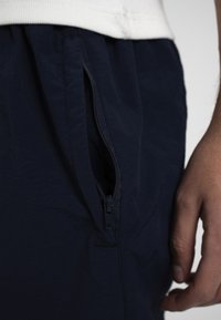 Gros plan d'un pantalon bleu marine avec une poche zippée ouverte à l'avant à côté d'une main posée de manière décontractée sur un fond blanc.