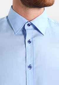 Lyseblå skjorte med klassisk krage, med fem knapper foran, inkludert to blå knapper og en glatt stoffoverflate.