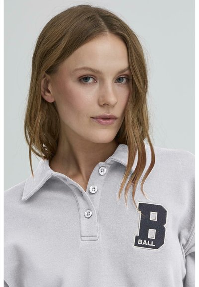 Svijetlosivi polo majica s kragnom, tri bijela gumba i crnim izvijenim dizajnom "B BALL" na prsima. Mekana tekstura.