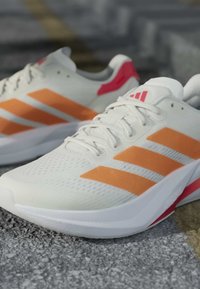 Chaussures de course blanches avec des bandes orange et des accents rouges posées sur une surface grise texturée, inclinées pour montrer le côté et l'avant.