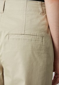 Nahaufnahme einer beigen Hose mit einer Gesäßtasche und Nähdetails, wobei ein Teil des Arms einer Person auf der rechten Seite sichtbar ist.