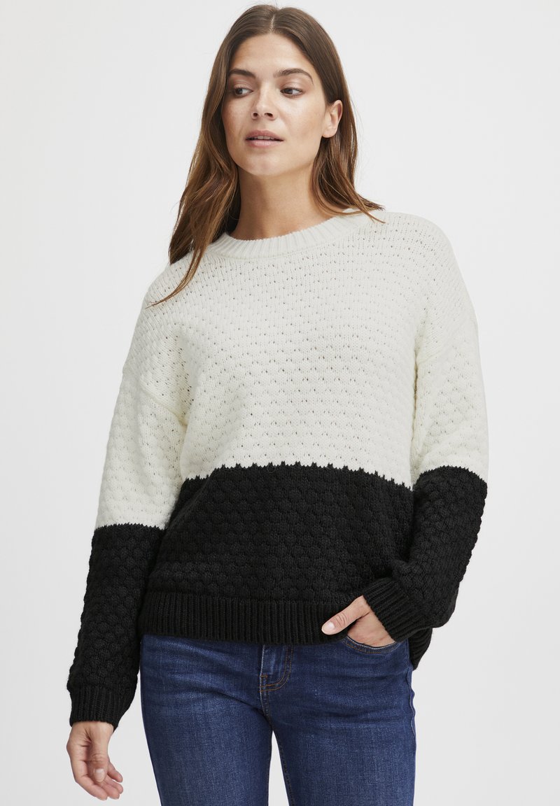 Pulover tricotat cu o secțiune superioară de culoare crem și o secțiune inferioară texturată de culoare neagră. Are un gât, manșete și tiv cu model în bulă. Are o croială lejeră, casual.