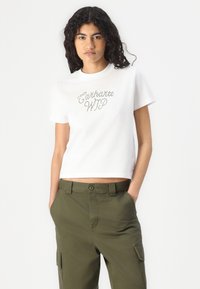 Weißes Baumwoll-T-Shirt mit kurzen Ärmeln, das "Carhartt WIP" in Strasssteinen auf der Brust trägt. Kombiniert mit olivgrünen Cargo-Hosen.