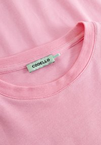 Roze katoenen T-shirt met een ronde halslijn, met een groene label waarop "CODELLO" staat, bevestigd aan de kraag. Glad materiaal, eenvoudig ontwerp.