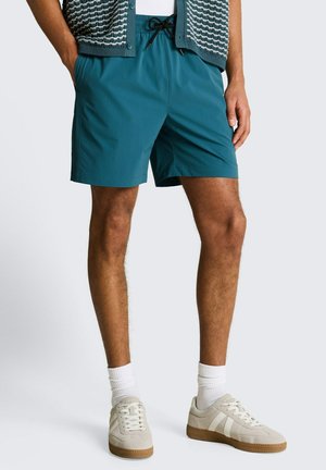 Uomo che indossa pantaloncini turchesi con coulisse, calzini bianchi, sneakers beige e un gilet di maglia blu sopra una camicia bianca, in piedi con le mani nelle tasche.