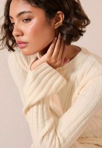 Vrouw met kort golvend haar, gekleed in een crèmekleurige geribbelde trui, die rustig haar kin op haar hand steunt met een kalme, bedachtzame uitdrukking.