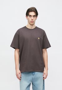 Νέος άνδρας με ίσια καστανά μαλλιά, ντυμένος με σκούρο καστανό T-shirt Carhartt και ανοιχτόχρωμο μπλε τζιν, στέκεται μπροστά σε ένα απλό λευκό φόντο.