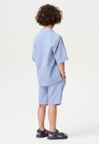 Ensemble composé d'une chemise à manches courtes et d'un short en motif vichy bleu, fabriqué dans un tissu léger. Comprend des poches latérales et des sandales décontractées.