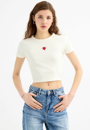 Camiseta corta blanca con un gráfico de un corazón rojo en el centro. Combinada con unos jeans de mezclilla azul claro de cintura alta.