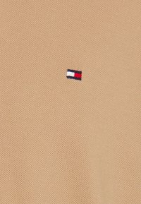 Närbild av tan stickad tyg med en liten broderad logotyp med röd, vit och blå rand i nedre vänster hörn.